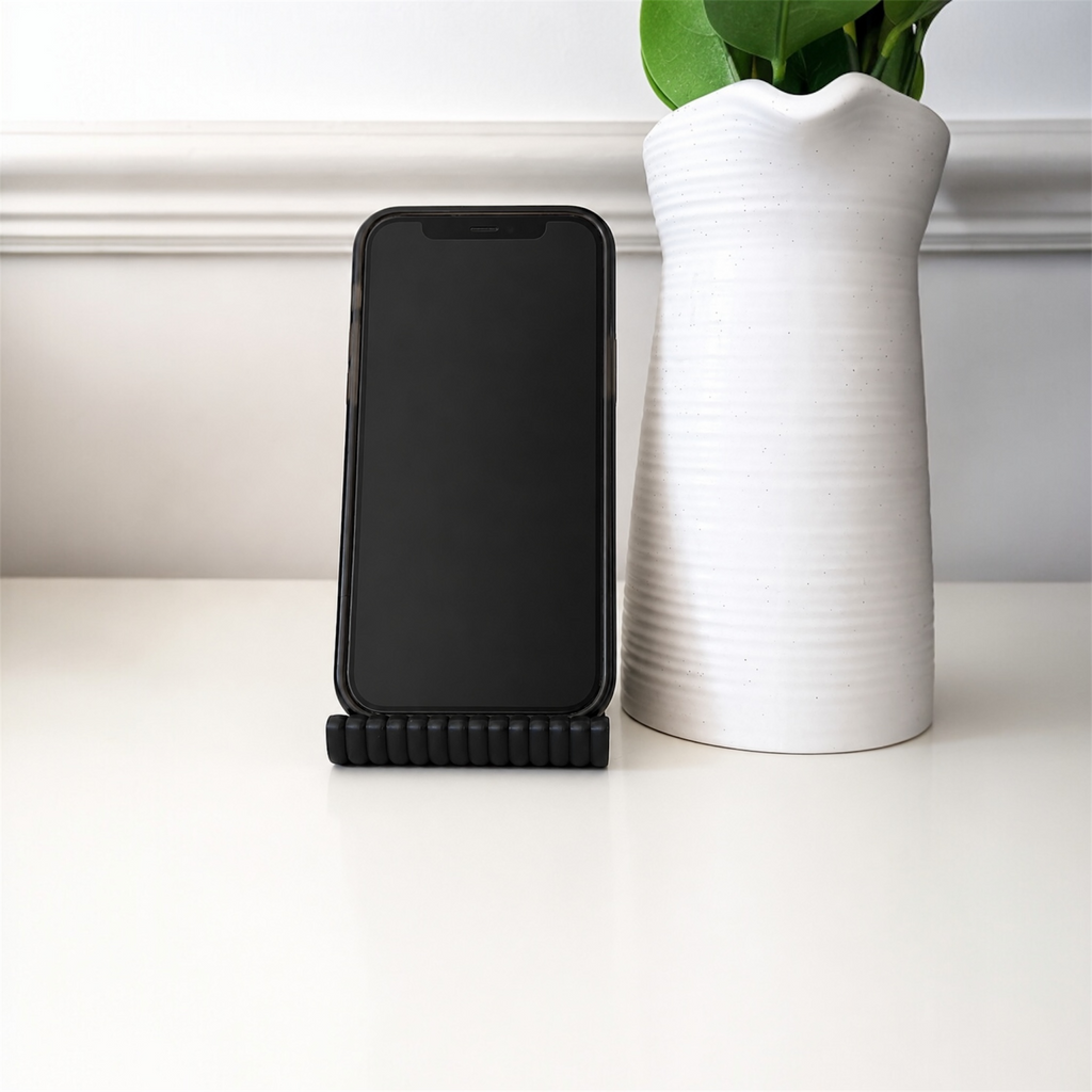Black phone stand with a smartphone on a white surface next to a white vase with green leaves.